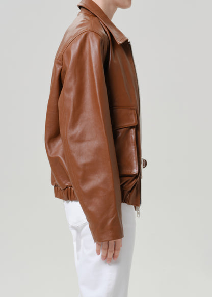 A side profile view of a person wearing a brown leather jacket and white pants against a light gray studio background.