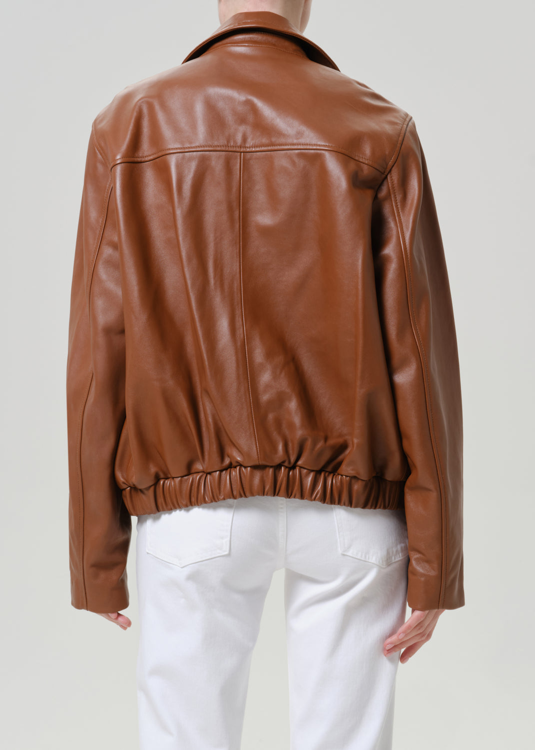 A person shown from the back wearing a brown leather jacket with a gathered elastic hem, paired with white pants, against a light gray studio background.