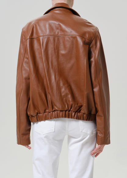 A person shown from the back wearing a brown leather jacket with a gathered elastic hem, paired with white pants, against a light gray studio background.