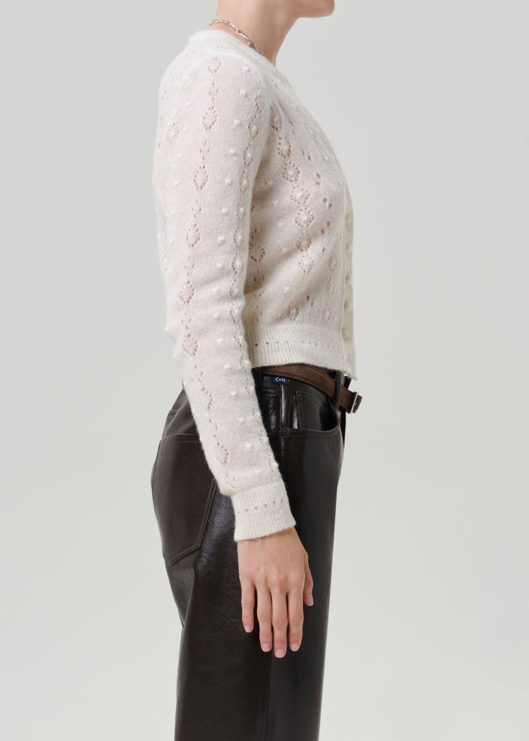 Woman wearing an ivory button-front cardigan with black leather pants viewed from the side on a grey background.