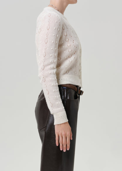 Woman wearing an ivory button-front cardigan with black leather pants viewed from the side on a grey background.