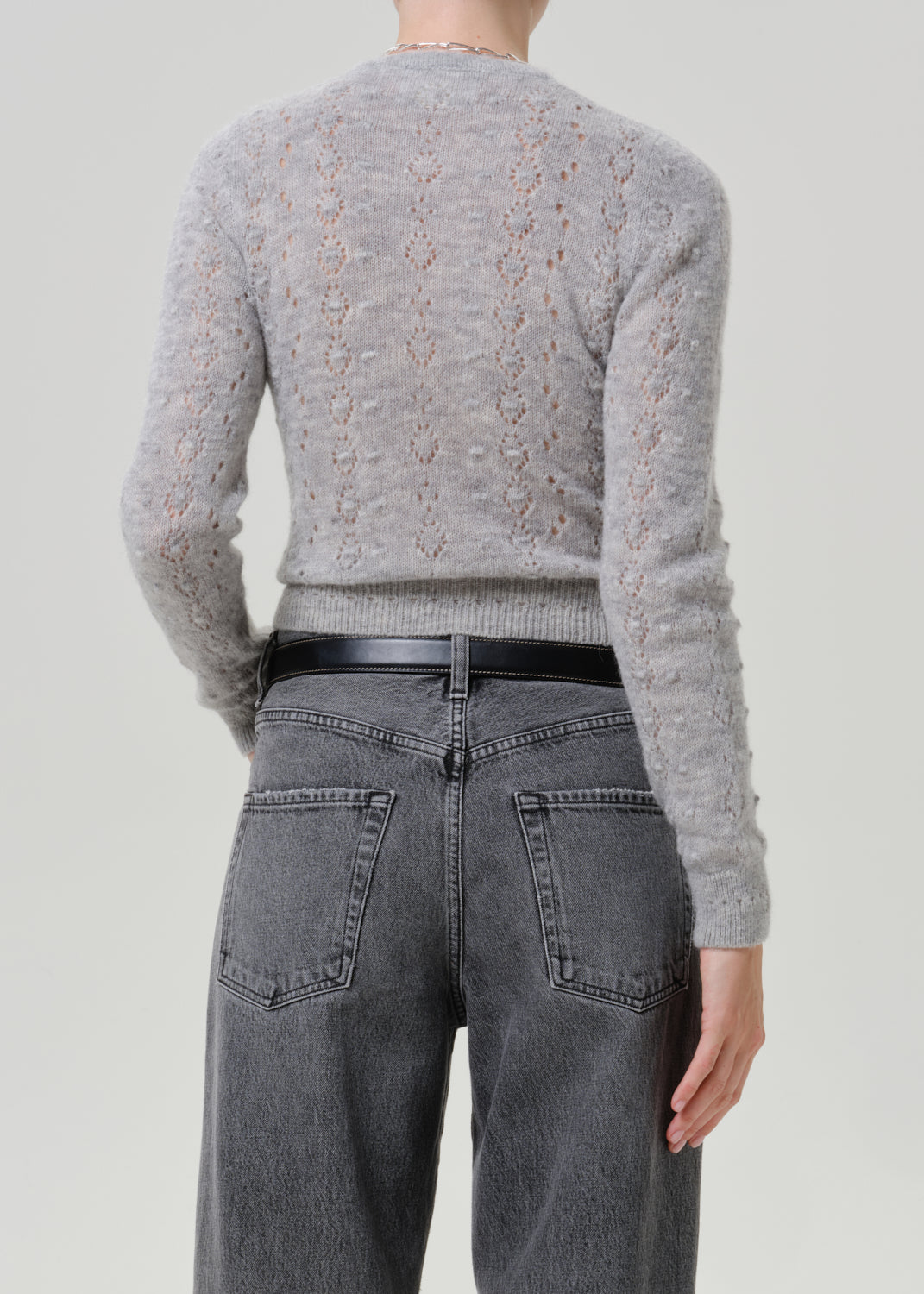 Woman wearing a grey button-front cardigan with dark grey jeans viewed from the back on a grey background.