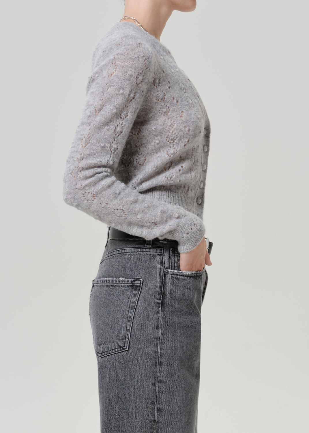 Woman wearing a grey button-front cardigan with dark grey jeans viewed from the side on a grey background.