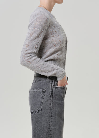 Woman wearing a grey button-front cardigan with dark grey jeans viewed from the side on a grey background.