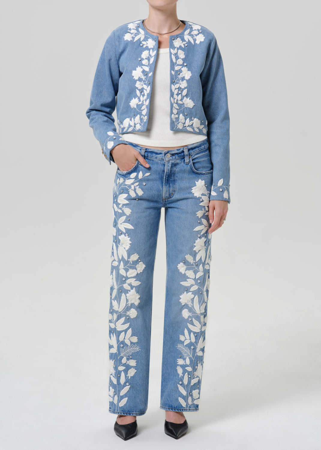 Woman wearing a light blue floral denim jacket with matching light blue floral jeans on a grey background.