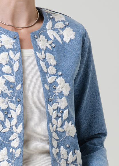 Close-up of a light blue floral denim jacket on a grey background.