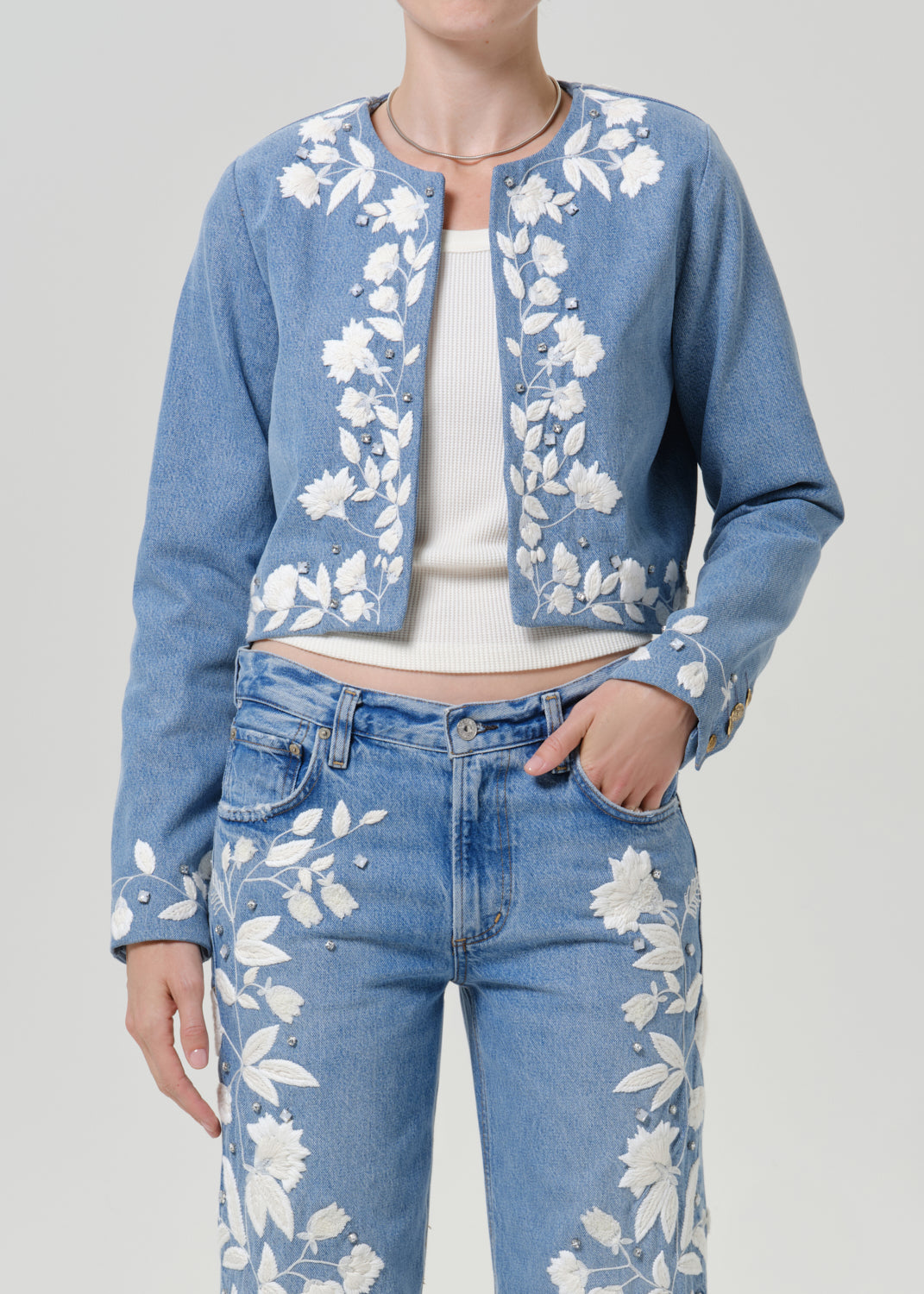 Woman wearing a light blue floral denim jacket with matching light blue floral jeans on a grey background.