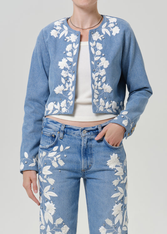 Woman wearing a light blue floral denim jacket with matching light blue floral jeans on a grey background.