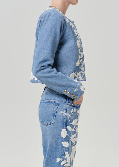 Woman wearing a light blue floral denim jacket with matching light blue floral jeans viewed from the side on a grey background.