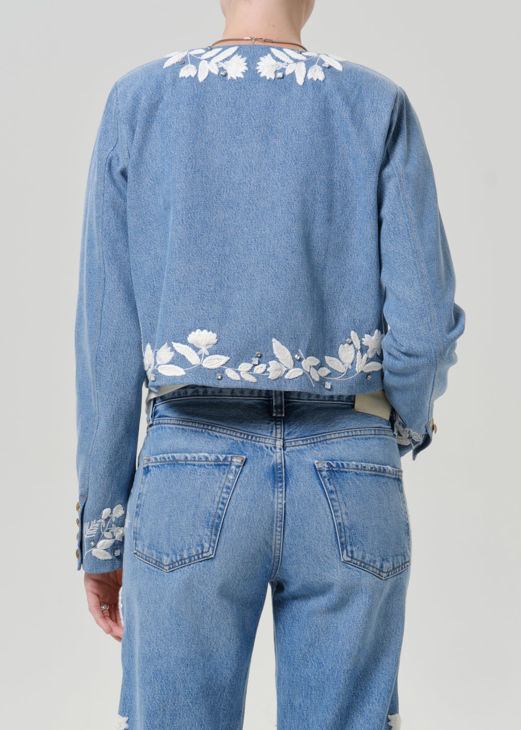 Woman wearing a light blue floral denim jacket with matching light blue floral jeans viewed from the back on a grey background.