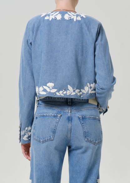 Woman wearing a light blue floral denim jacket with matching light blue floral jeans viewed from the back on a grey background.
