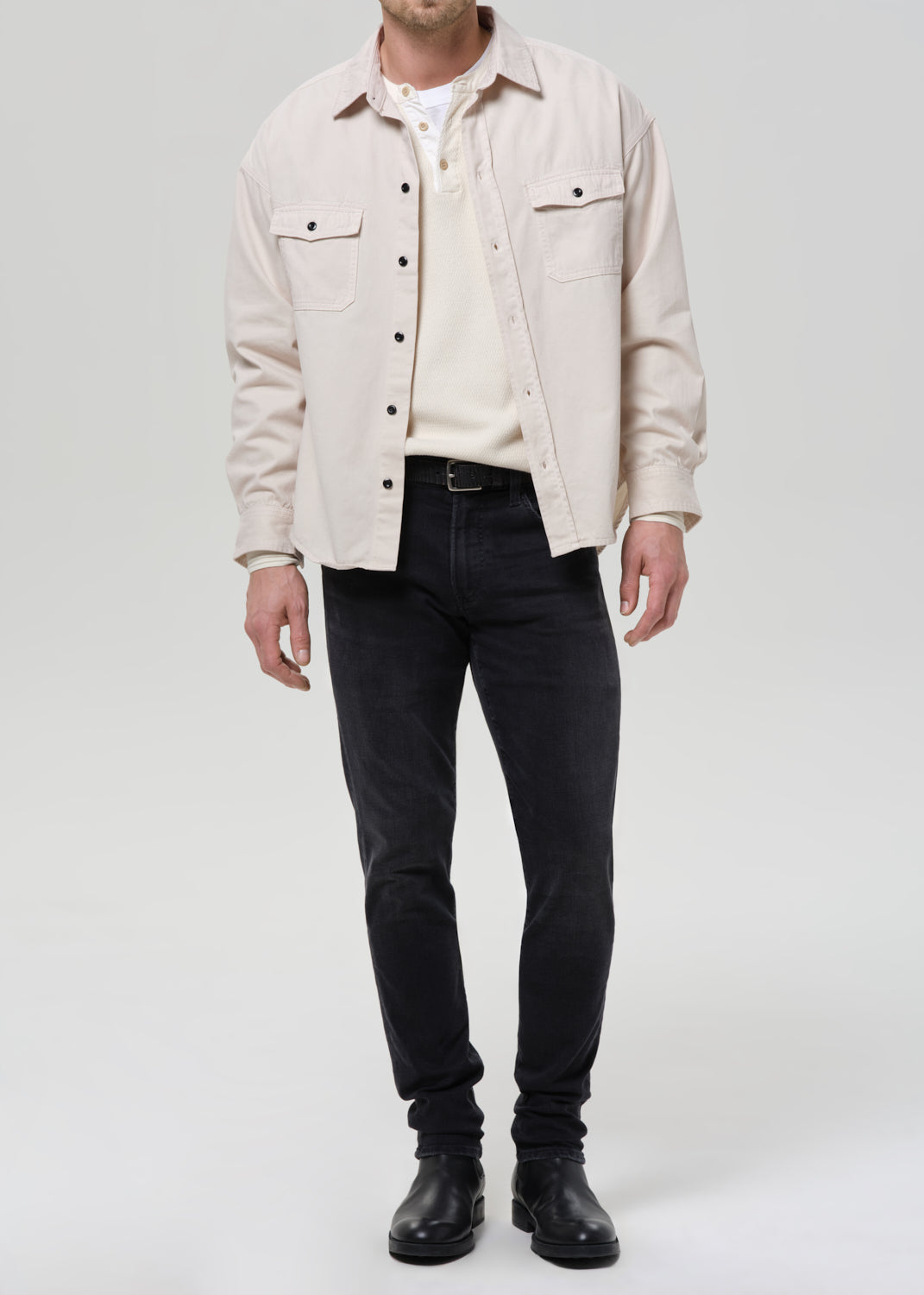 A man wearing a light beige overshirt over a cream henley shirt, paired with dark charcoal jeans and black leather boots, standing against a grey background.