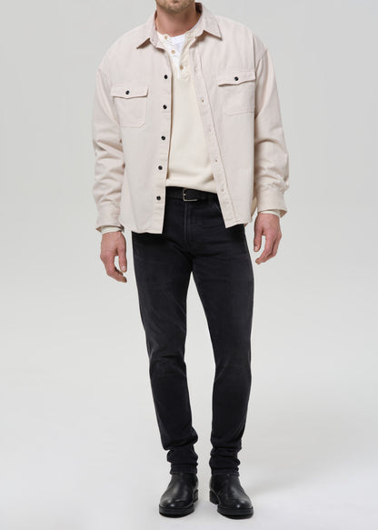 A man wearing a light beige overshirt over a cream henley shirt, paired with dark charcoal jeans and black leather boots, standing against a grey background.