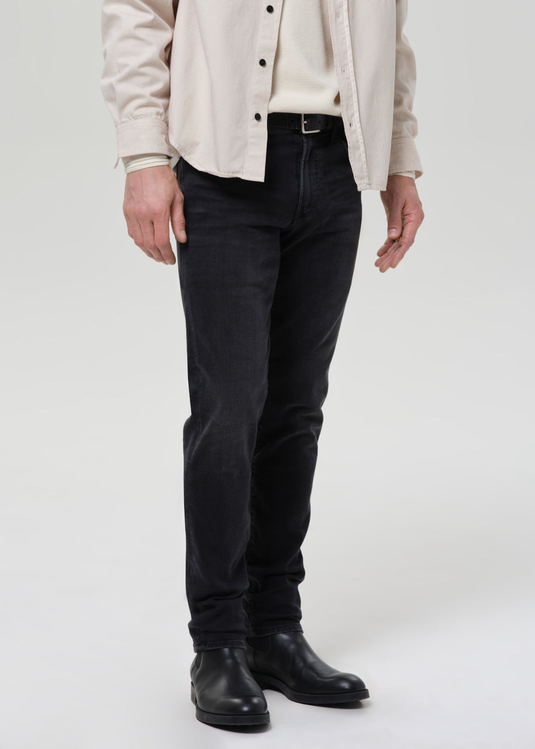 A person wearing slim-fit black jeans, a cream shirt, a light beige overshirt, and black leather boots, shown from the chest down against a grey background.