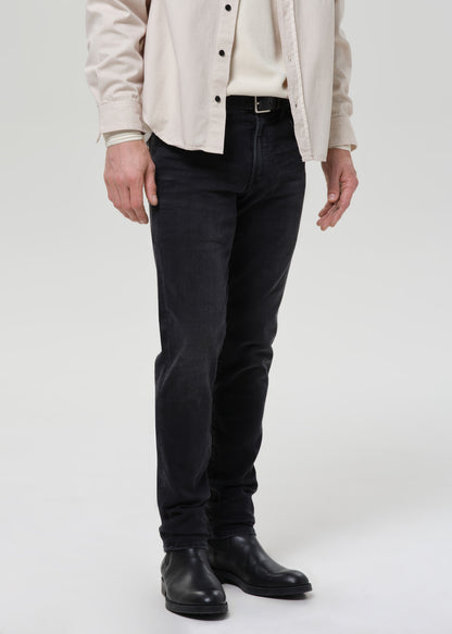 A person wearing slim-fit black jeans, a cream shirt, a light beige overshirt, and black leather boots, shown from the chest down against a grey background.