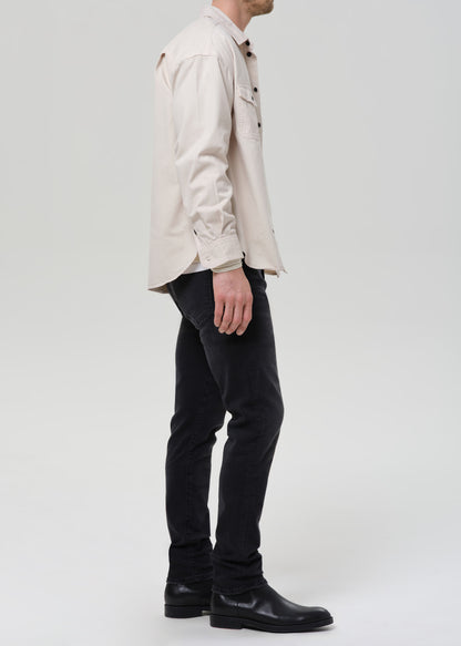 A man shown in profile wearing a light beige long-sleeve button-up shirt, black jeans, and black leather boots, photographed against a grey background.
