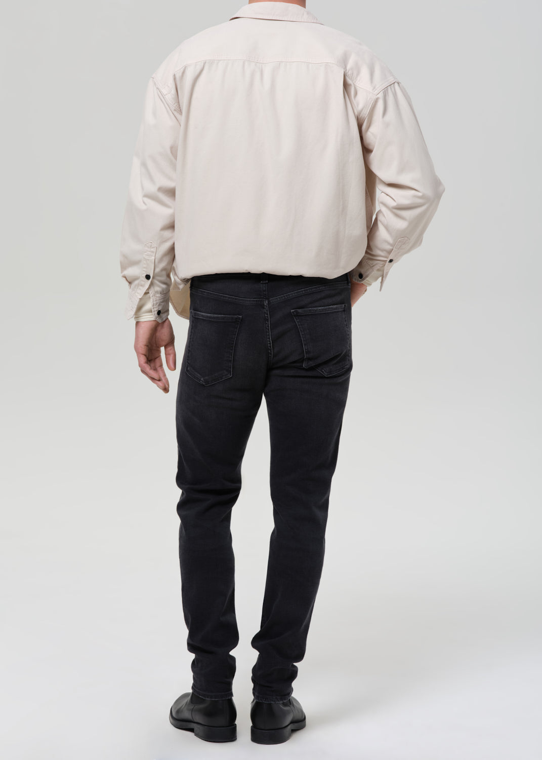 A man shown from the back wearing a light beige oversized shirt, dark charcoal slim-fit jeans, and black boots, photographed against a grey background.