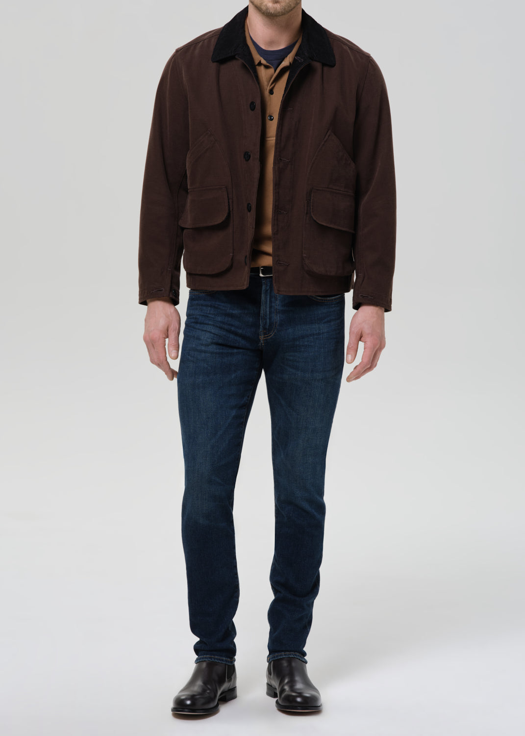 Person wearing dark indigo straight leg jeans with a brown chore jacket and brown crewneck tee on a grey background.