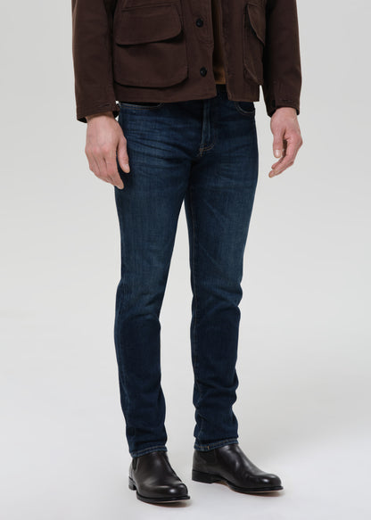 Person wearing dark indigo straight leg jeans with a brown chore jacket and brown crewneck tee on a grey background.