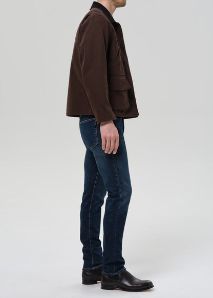 Person wearing dark indigo straight leg jeans with a brown chore jacket and brown crewneck tee viewed from the side on a grey background.