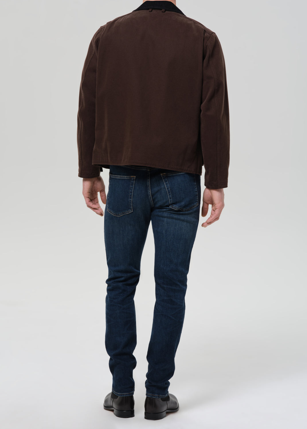 Person wearing dark indigo straight leg jeans with a brown chore jacket and brown crewneck tee viewed from the back on a grey background.