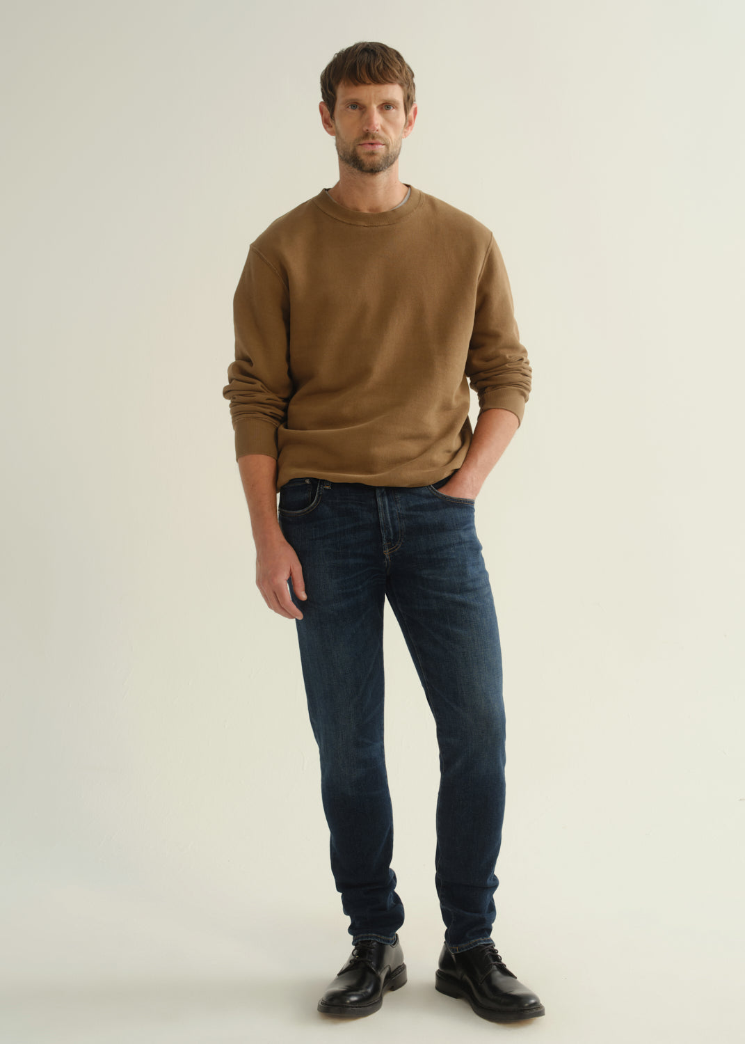 Person wearing dark indigo slim tapered jeans with a brown crewneck sweatshirt and black shoes, front view.
