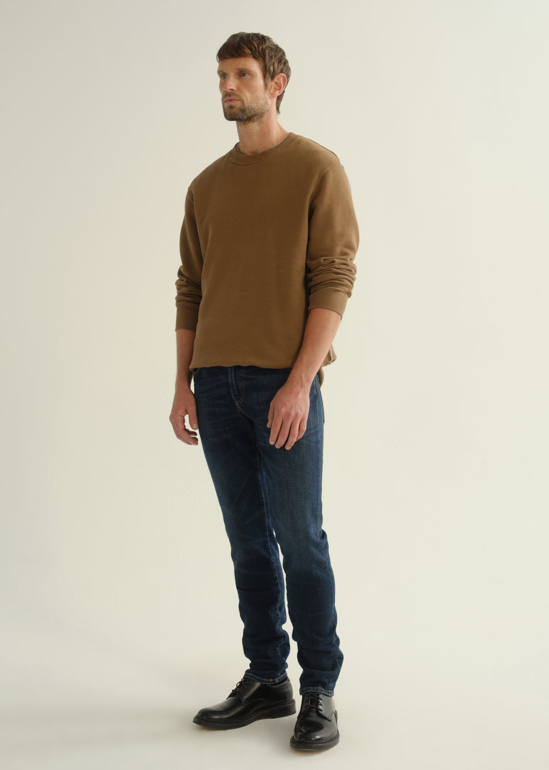 Person wearing dark indigo slim tapered jeans with a brown crewneck sweatshirt and black shoes, front angled view.