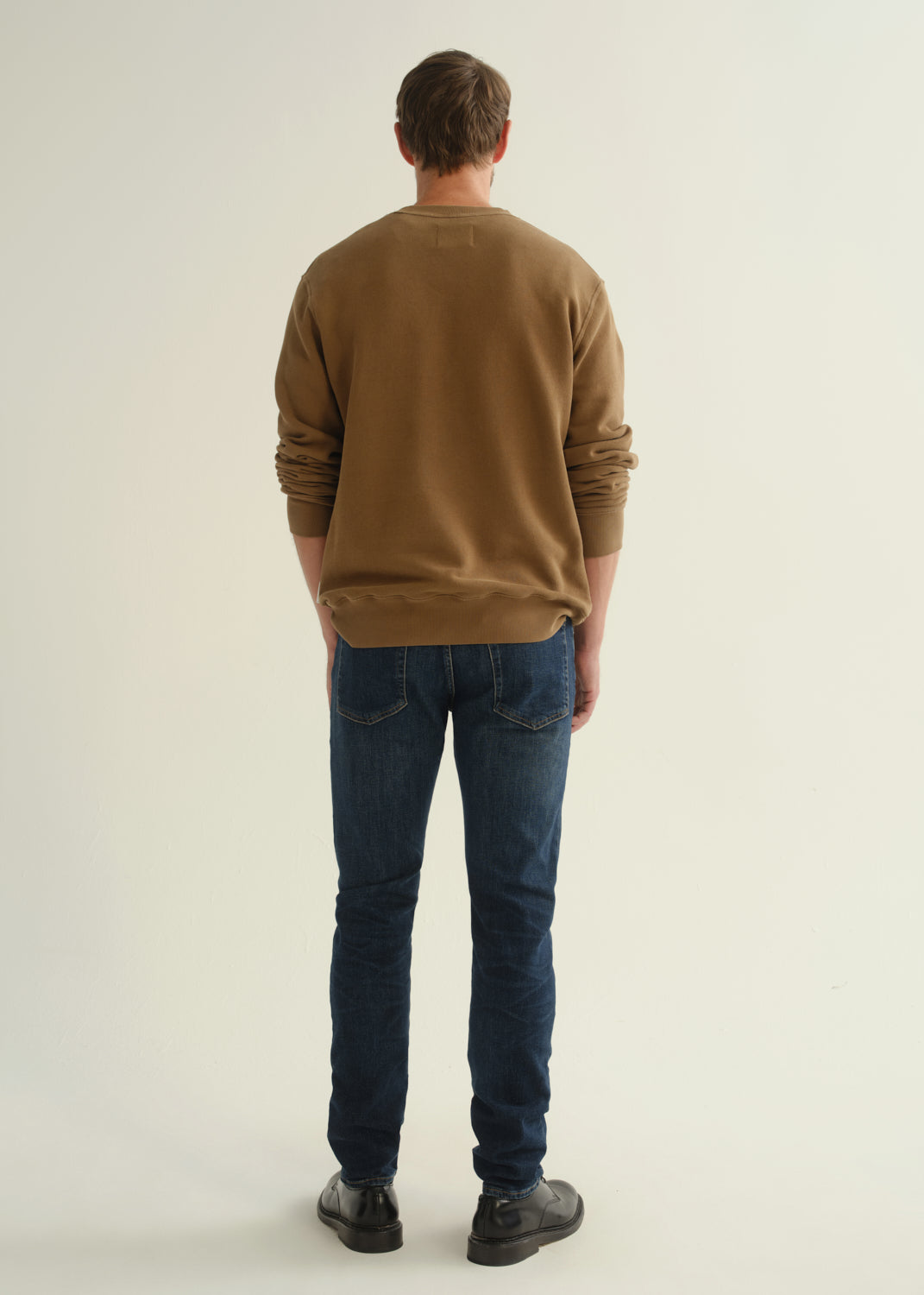 Person wearing dark indigo slim tapered jeans with a brown crewneck sweatshirt and black shoes, front angled view.