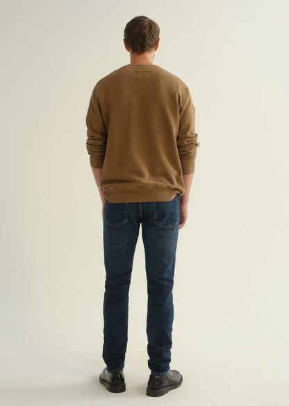 Person wearing dark indigo slim tapered jeans with a brown crewneck sweatshirt and black shoes, front angled view.