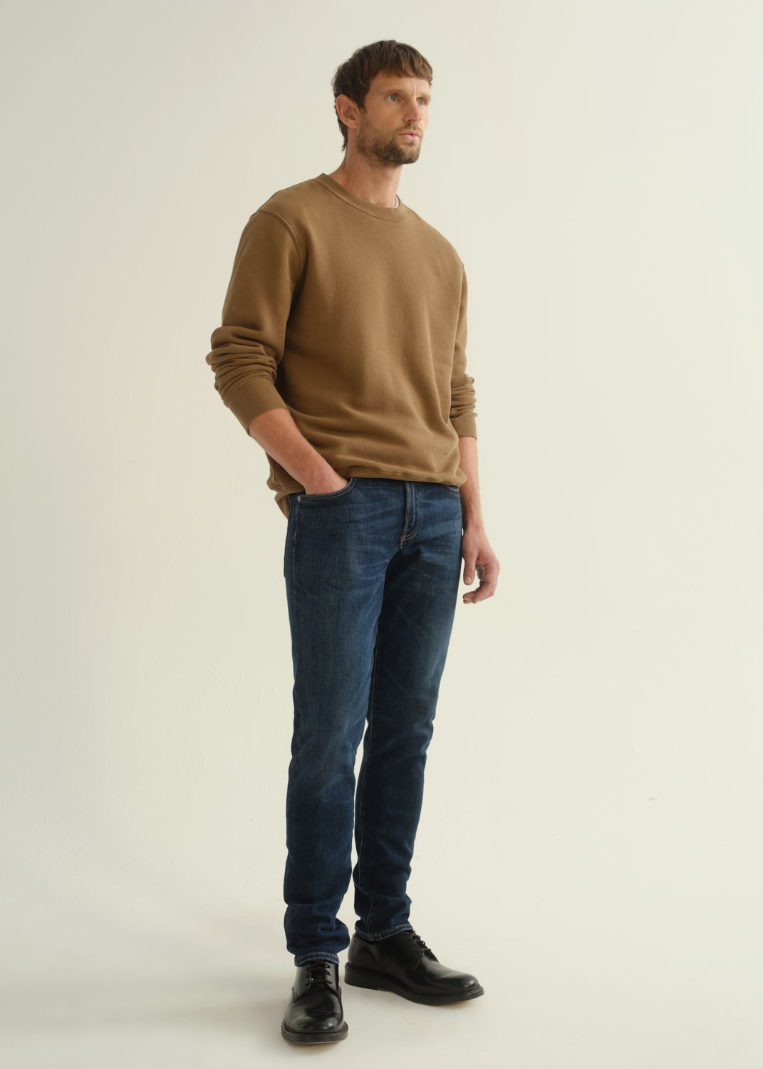 Person wearing dark indigo slim tapered jeans with a brown crewneck sweatshirt and black shoes, back view.