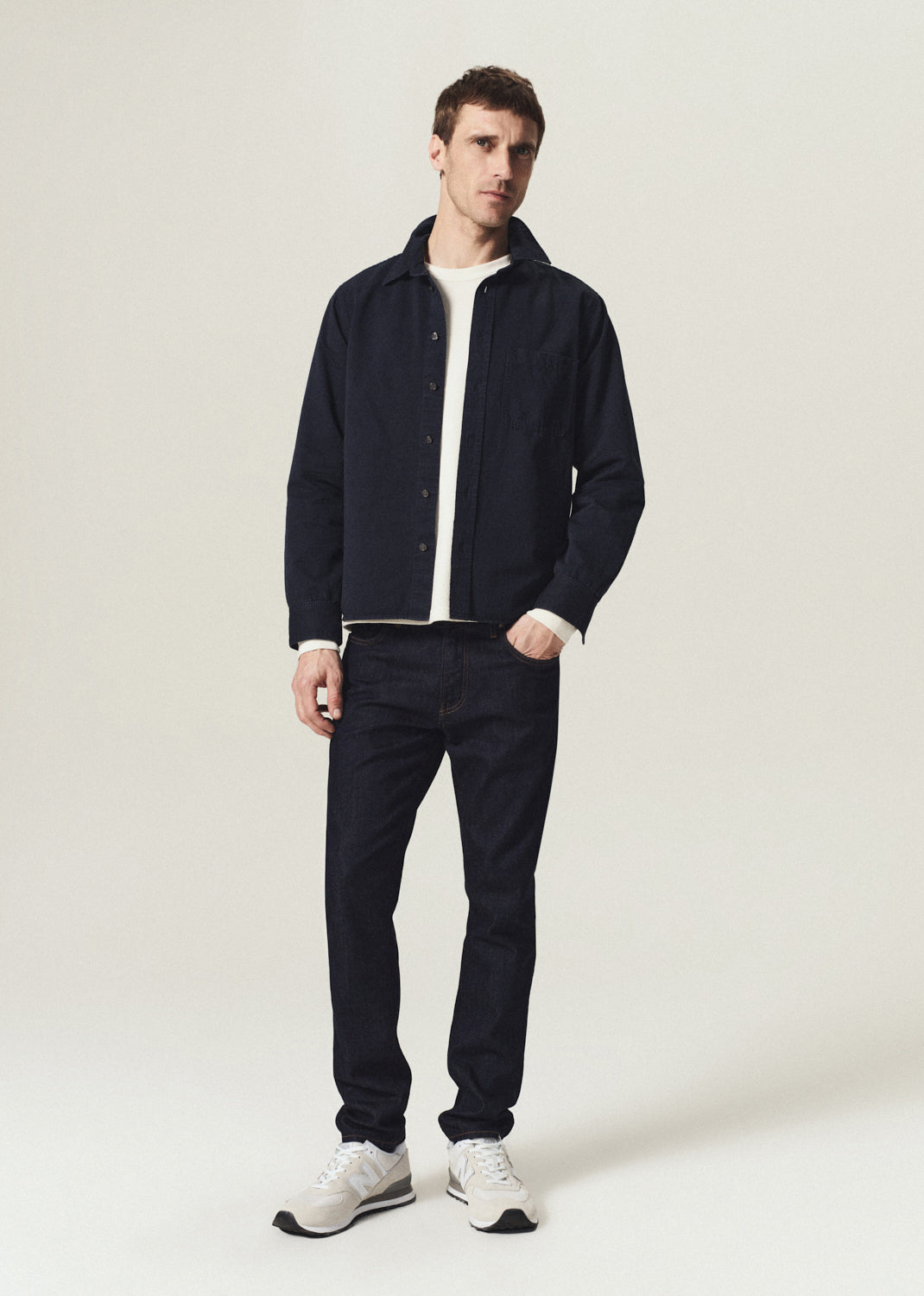 A man modeling a dark navy blue overshirt jacket over a white t-shirt, paired with dark navy jeans and white sneakers, standing against a light beige studio background.