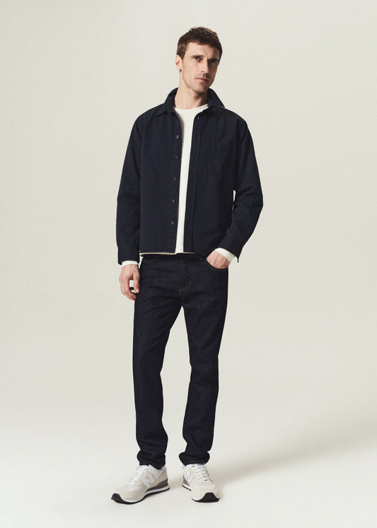 A man modeling a dark navy blue overshirt jacket over a white t-shirt, paired with dark navy jeans and white sneakers, standing against a light beige studio background.
