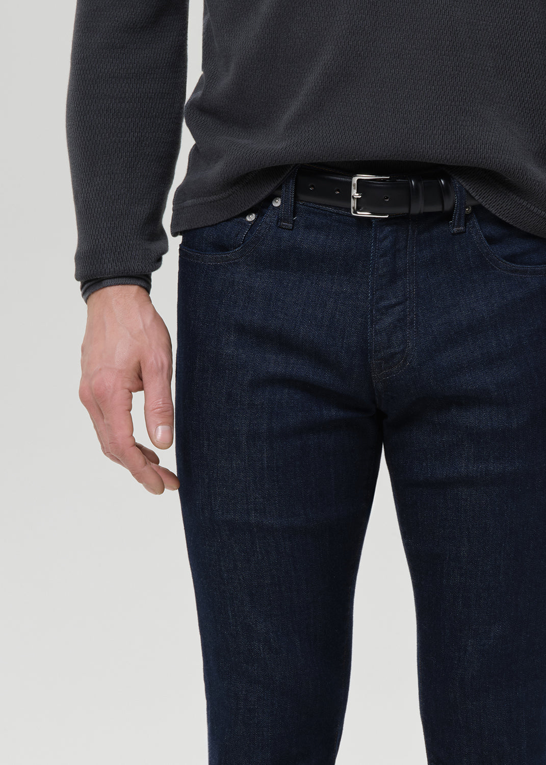 A close-up of a person wearing dark blue slim-fit jeans with a black belt, paired with a dark charcoal ribbed long-sleeve top, against a light gray background.