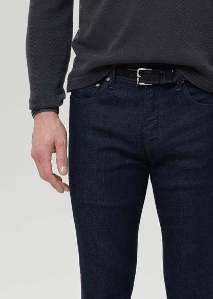 A close-up of a person wearing dark blue slim-fit jeans with a black belt, paired with a dark charcoal ribbed long-sleeve top, against a light gray background.