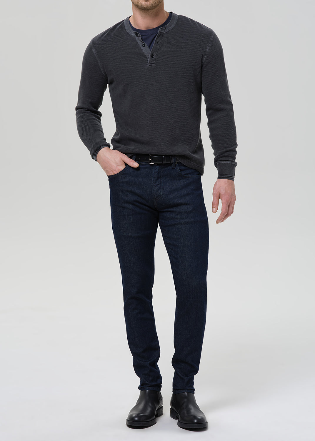 A man wearing a dark charcoal gray long-sleeve henley shirt, dark blue slim-fit jeans, and black leather boots, standing against a light gray studio background.