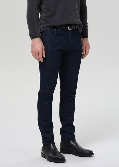 A person wearing slim-fit dark blue jeans, a dark charcoal long-sleeve top, a black belt, and black leather boots, photographed against a light gray studio background.