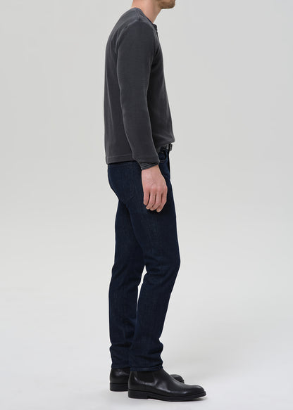 A man shown in profile wearing a dark charcoal long-sleeve shirt, dark blue jeans, and black leather boots against a light gray studio background.