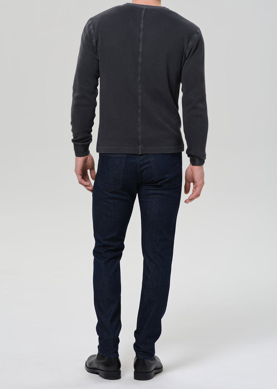 A man shown from the back wearing a dark charcoal gray long-sleeve crewneck sweater and dark blue jeans, standing against a light gray studio background.