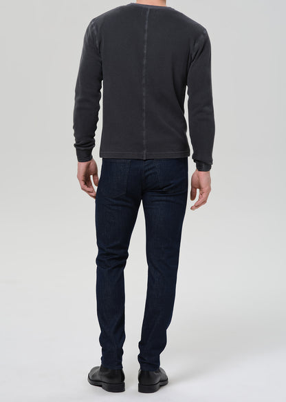A man shown from the back wearing a dark charcoal gray long-sleeve crewneck sweater and dark blue jeans, standing against a light gray studio background.