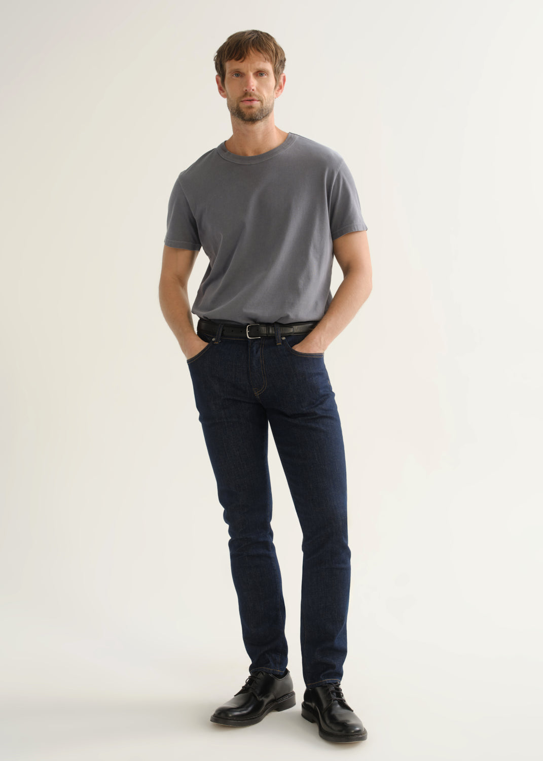 Person wearing dark indigo slim tapered jeans with a grey short sleeve crewneck t-shirt and black shoes, front view.