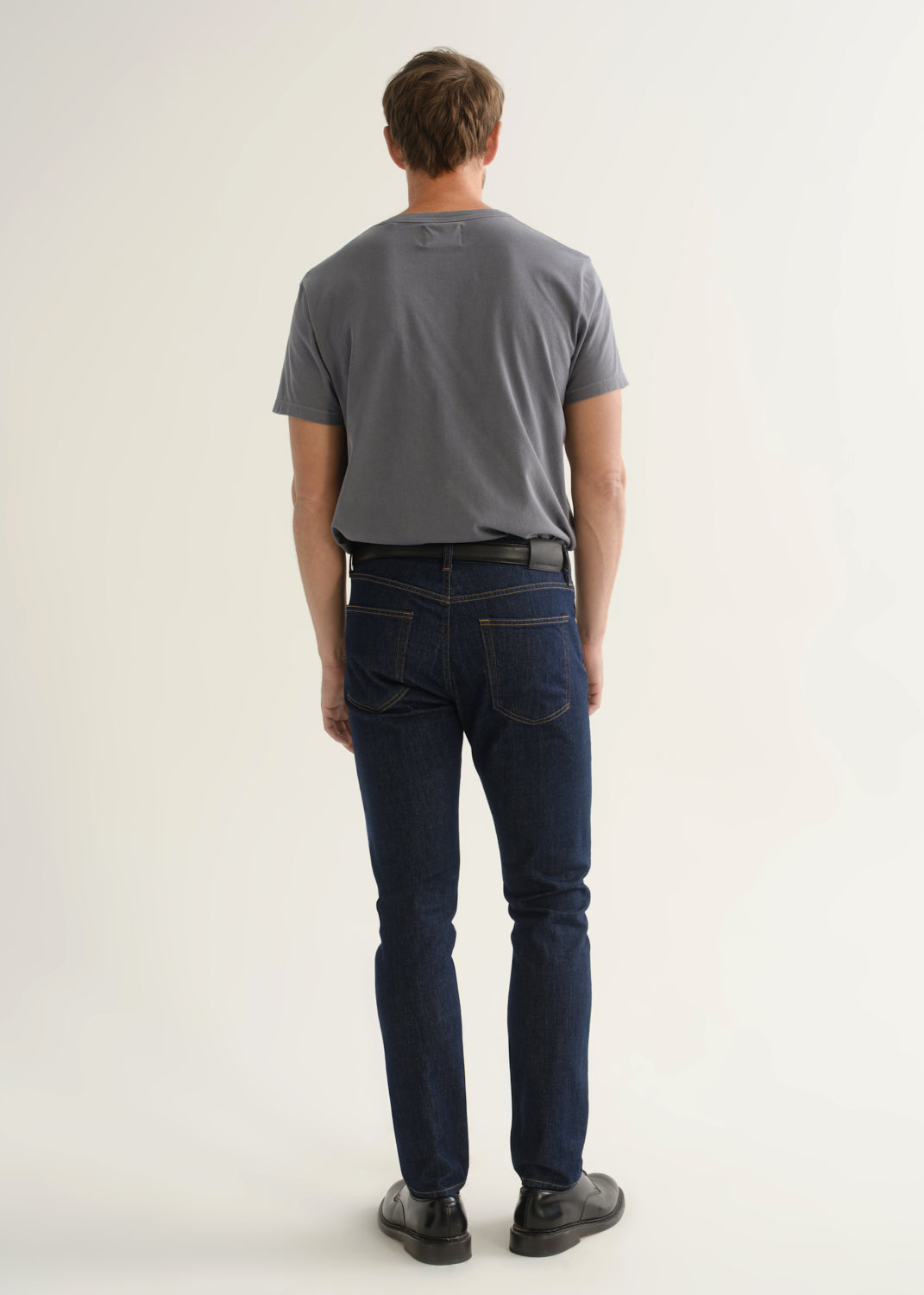 Person wearing dark indigo slim tapered jeans with a grey short sleeve crewneck t-shirt and black shoes, back view.