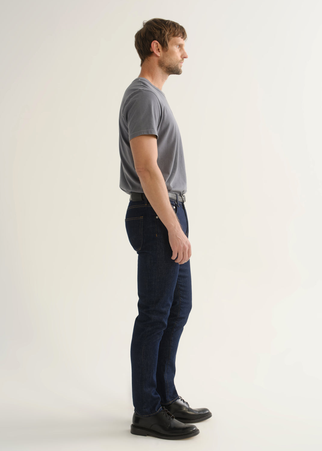 Person wearing dark indigo slim tapered jeans with a grey short sleeve crewneck t-shirt and black shoes, side view.