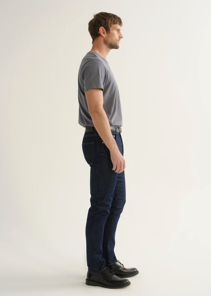 Person wearing dark indigo slim tapered jeans with a grey short sleeve crewneck t-shirt and black shoes, side view.
