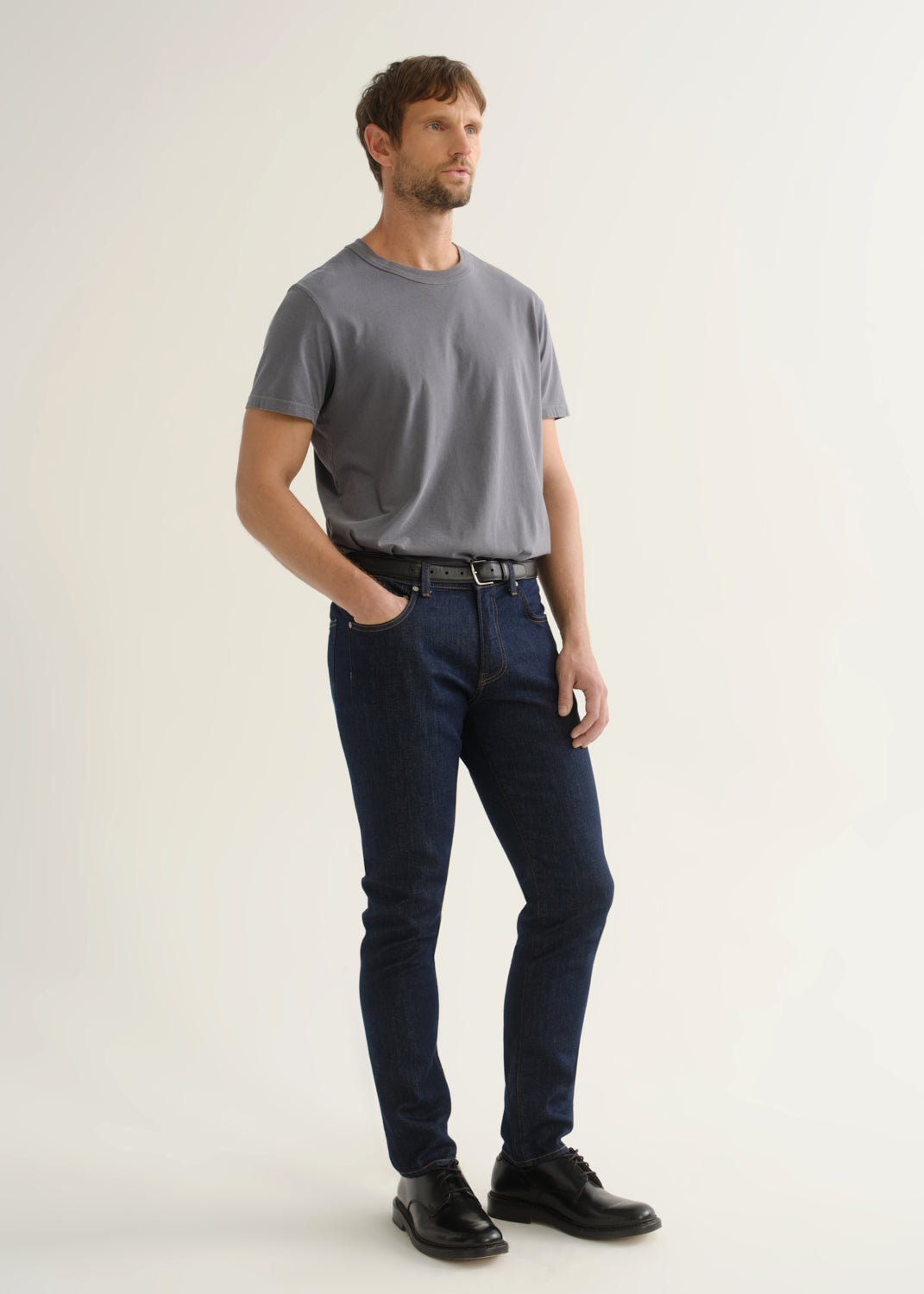 Person wearing dark indigo slim tapered jeans with a grey short sleeve crewneck t-shirt and black shoes, side view.