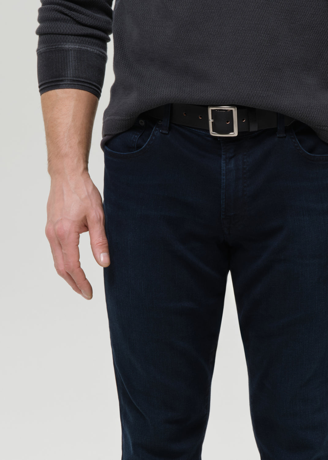 Close-up of a person wearing dark navy blue jeans, a black belt with a silver buckle, and a dark charcoal ribbed sweater against a grey background.