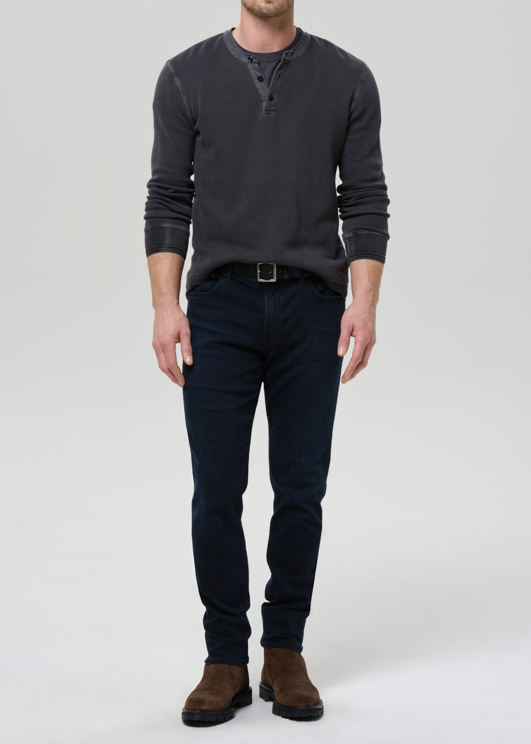 A man modeling a dark charcoal grey  long-sleeve henley shirt paired with dark navy blue jeans and brown suede boots, standing against a grey background.