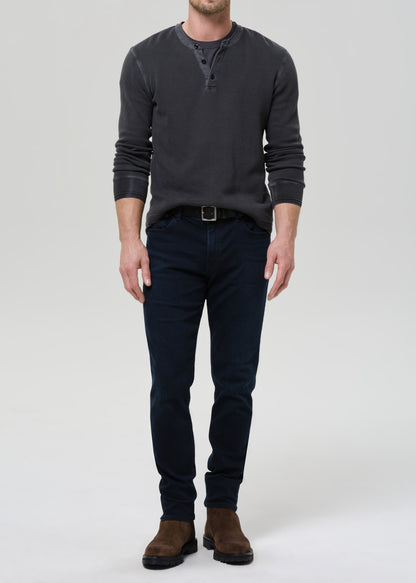A man modeling a dark charcoal grey  long-sleeve henley shirt paired with dark navy blue jeans and brown suede boots, standing against a grey background.