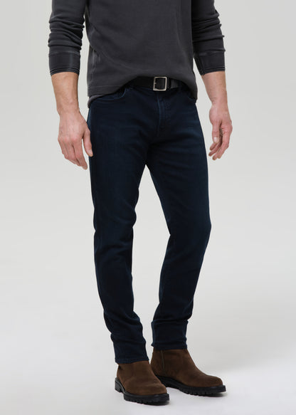 A man wearing slim-fit dark navy blue jeans, a dark grey long-sleeve shirt, a black belt, and brown suede ankle boots, standing against a grey background.