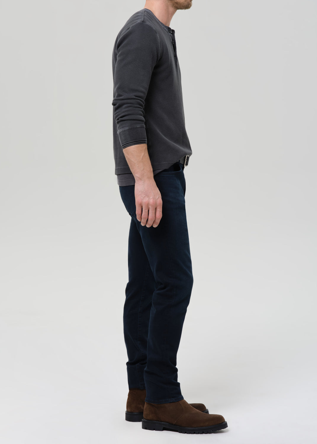 A man shown in profile wearing a dark charcoal grey  long-sleeve henley shirt, dark navy blue jeans, and brown suede boots, standing against a grey background.