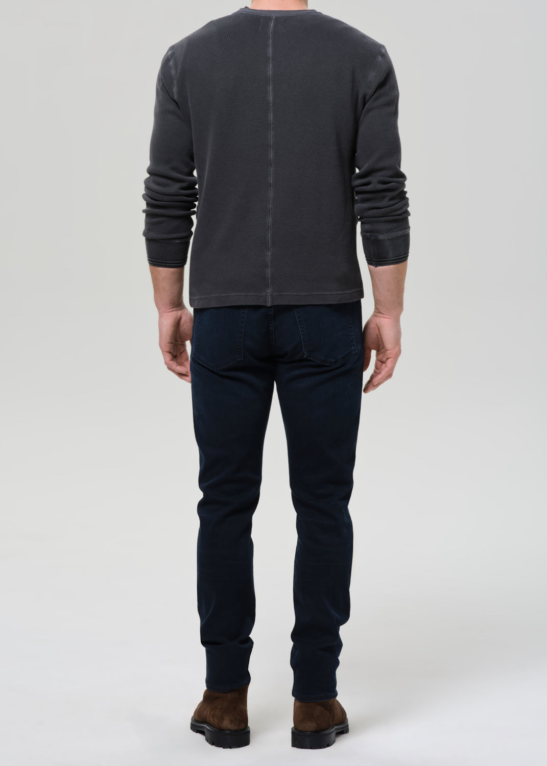 A man viewed from the back wearing a dark charcoal grey  long-sleeve crewneck shirt, dark navy blue jeans, and brown boots, standing against a grey  background.